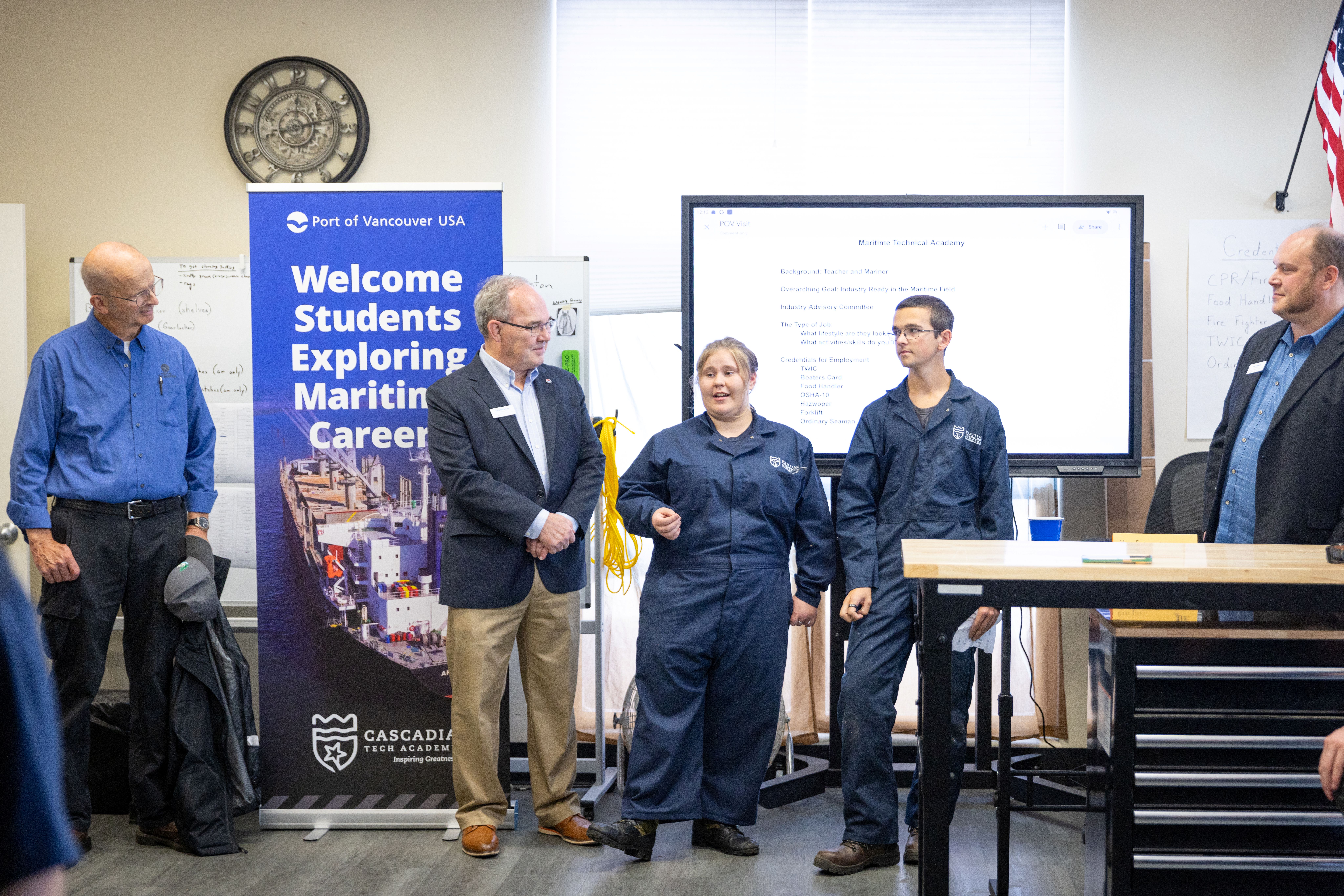 Students in maritime academy uniforms stand with port and school representatives during a presentation about maritime careers. A Port of Vancouver USA banner and presentation screen are visible in the background.