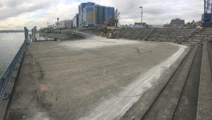 Concrete amphitheater with concrete steps sits along river's edge with tall buildings under construction in the background. 