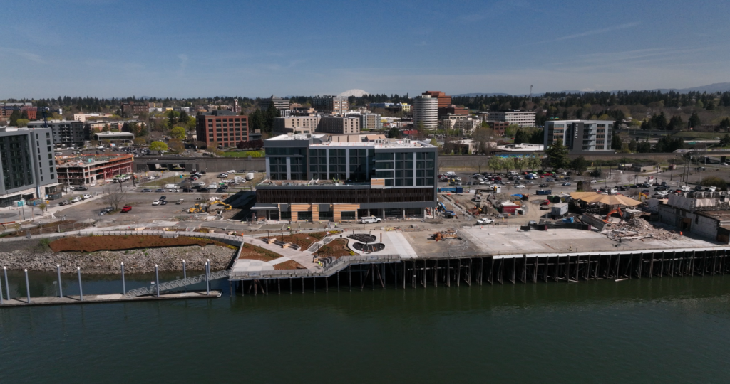 POV birds eye view in the Columbia River looking north at a long, aged dock. a single new high-rise building sits behind the dock on the shoreline. some fresh mulch dots the landscaping of a new dock area to the left of the aged dock. On the aged dock there is rubble from a torn down building. Blue skies with hints of clouds. Mt. Saint Helens can be seen in the distance in the middle of the photo.