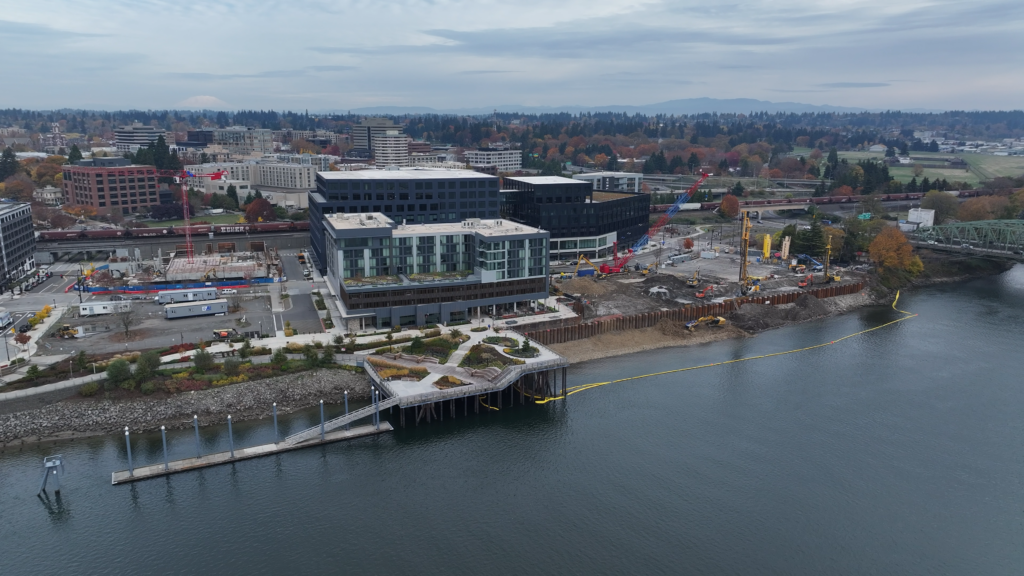 POV birds eye view in the Columbia River looking northeast at Terminal 1. A small dock with landscaping and pathways and a floating dock for boats sits in the center of the photo. To the east of that dock is an empty space where a dock used to be. Construction equipment is on the shoreline and along the riverbank. Three new high-rise buildings are on the shore behind the construction area. Cloudy skies with a very slight view of Mt. Saint Helens in the background.