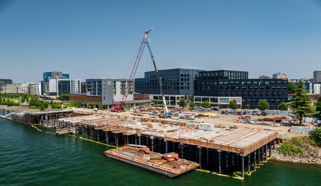 POV from middle of river looking north at the shoreline. A small barge with two lift booms floats in the river in front of a wide dock that is under construction. Two large cranes sit in the middle of the photo. Various construction materials and vehicles are on the shore in the construction area. Various high rise buildings create the backdrop to the photo. Blue skies with no clouds.