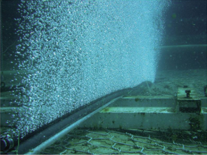 Underwater photo of a pipe along the bottom with a wall of bubbles rising from it.